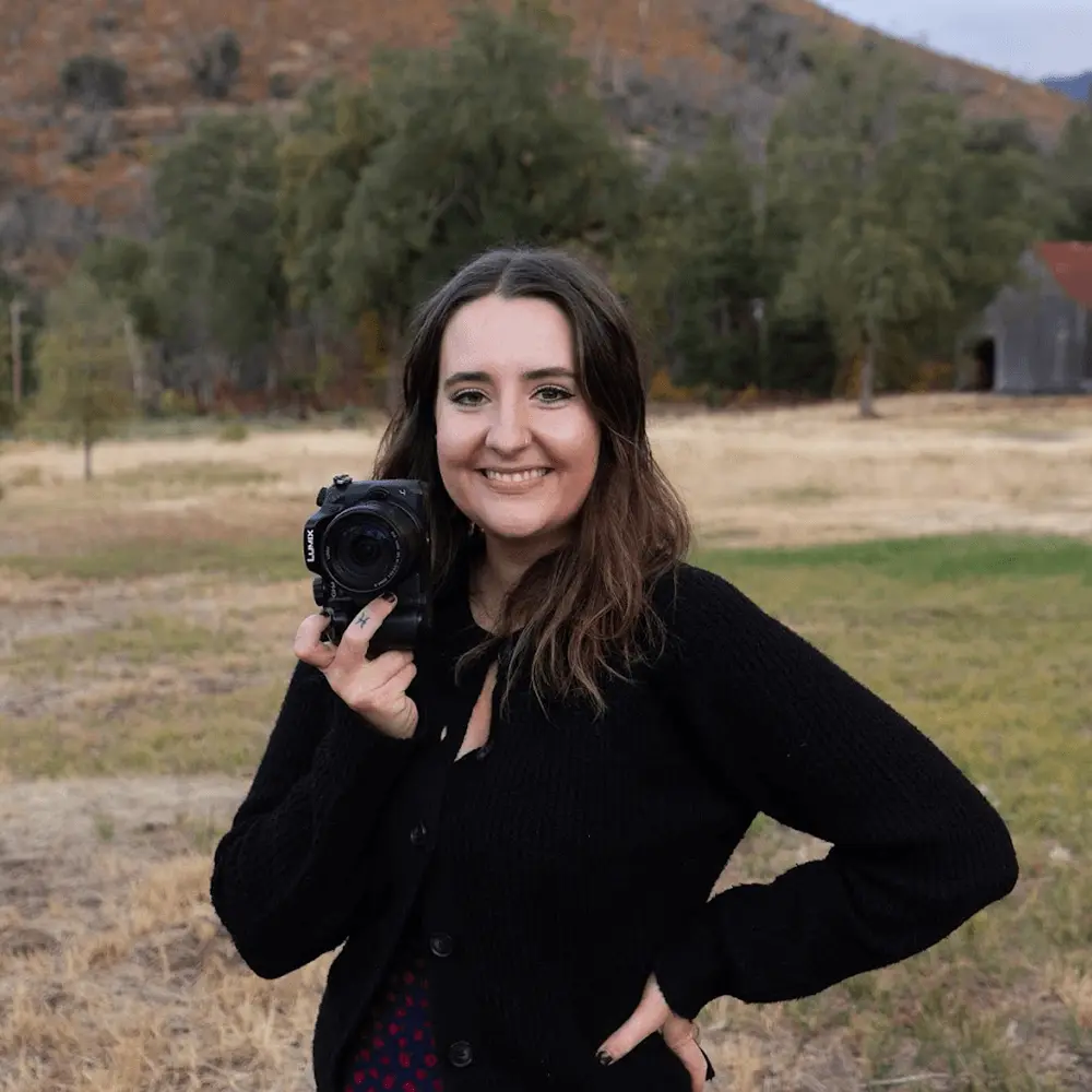 A white female photographer from Dallas-Fort Worth Texas smiling outside and posing with her professional camera.